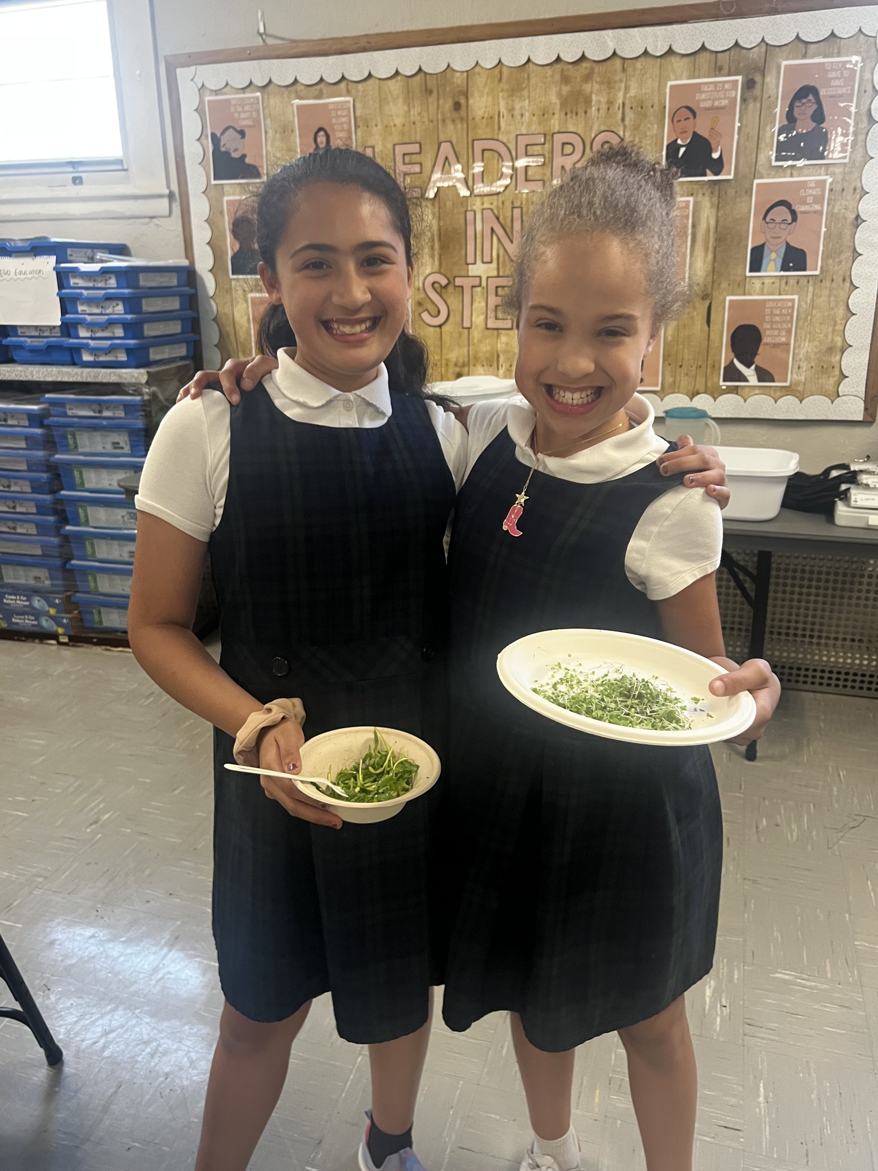 Two Boston students in school uniforms smiling and holding bowls of the microgreens they grew in class